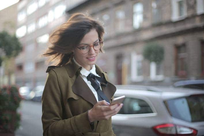 Meisje met een bril op loopt op straat en kijkt naar de telefoon in haar hand