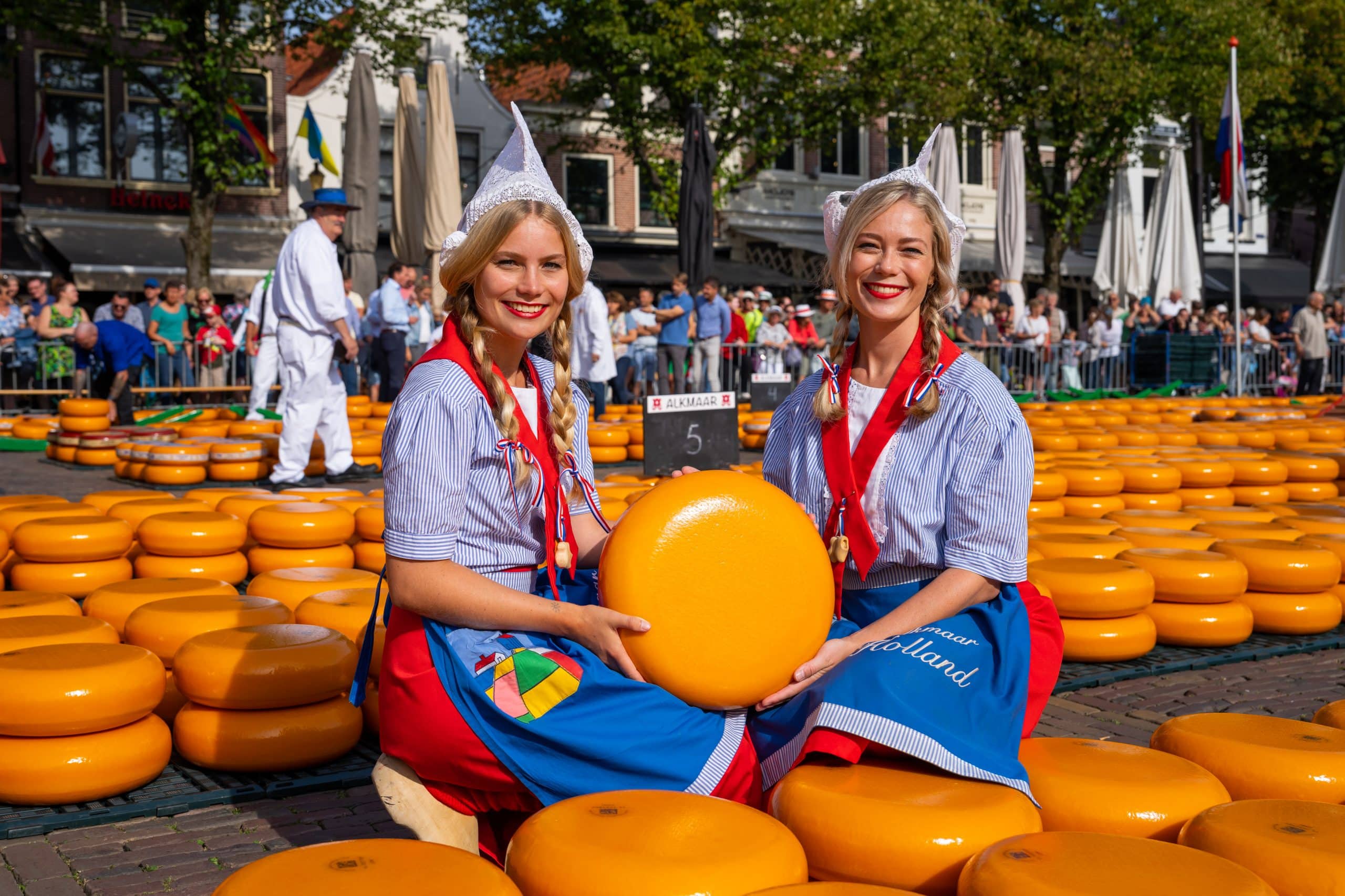 twee kaasmeisjes zitten op hun hurken en houden samen een kaas vast. Zij zijn op de kaasmarkt in Alkmaar.
