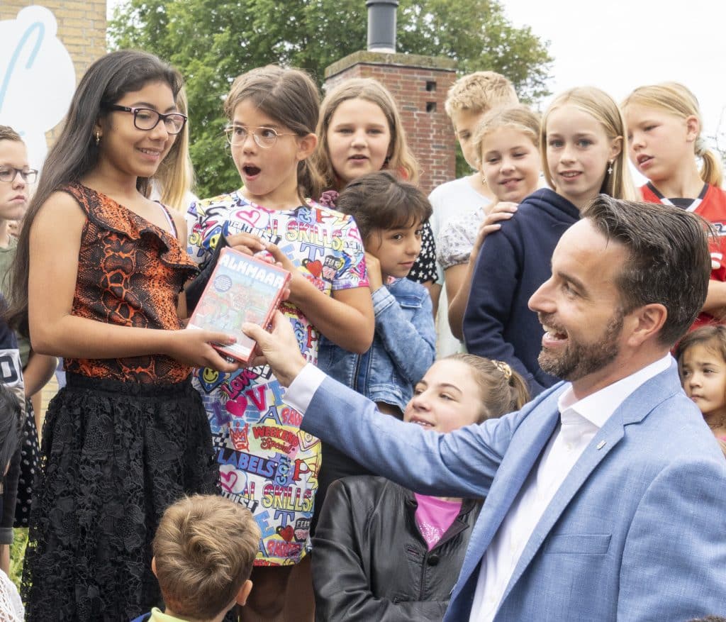 Malaika krijgt van wethouder Jasper Nieuwenhuizen een cadeautje overhandigd. Ze zijn op een schoolplein omringd met kinderen.