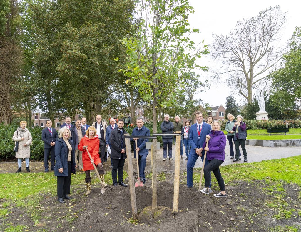 Groep mensen staan in een park. Een deel van de groep staat met een schep in de hand. Zij staan op het punt om een boom te planten. 