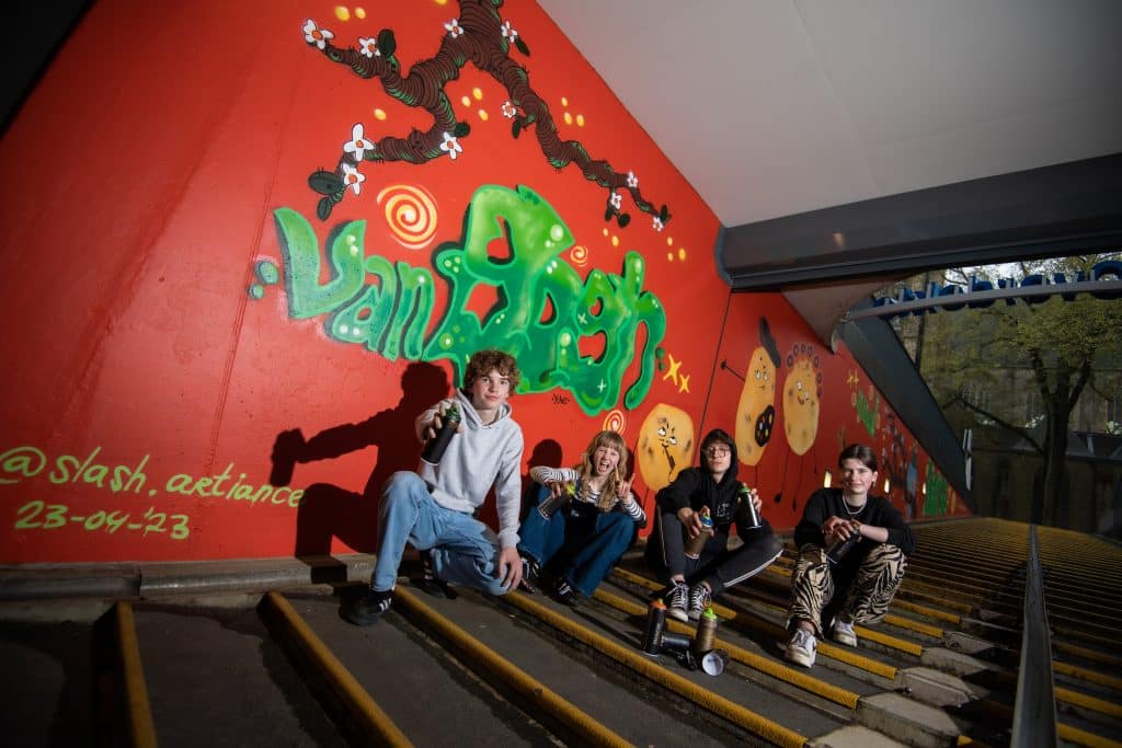 Foto van groep van vier jongeren op een trap. Achter hen is een wand met graffiti te zien.