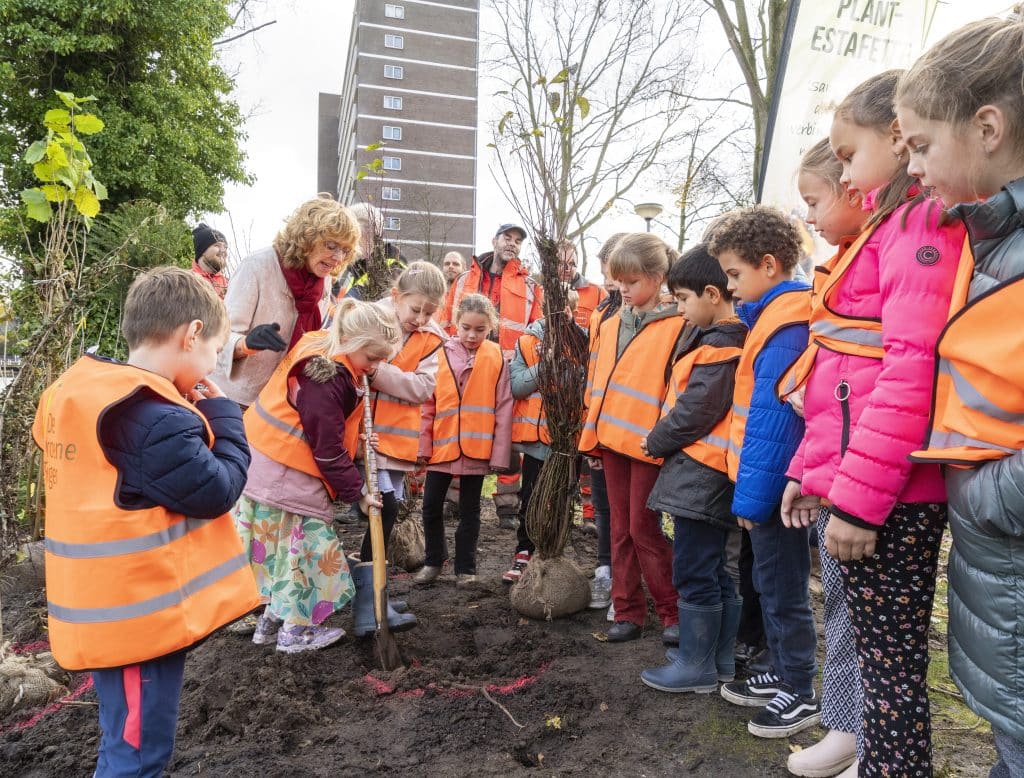 Een groep kinderen die samen met de wethouder een boompje planten met een spade. De kinderen dragen allemaal een oranje veiligheidshesje. 