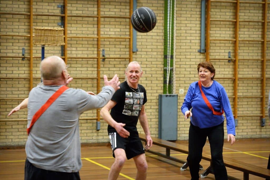 Op de foto zijn drie mensen aan het sporten in de gymzaal. Ze doen een balspel. Sporten helpt bij het vitaal en gezond ouder worden.