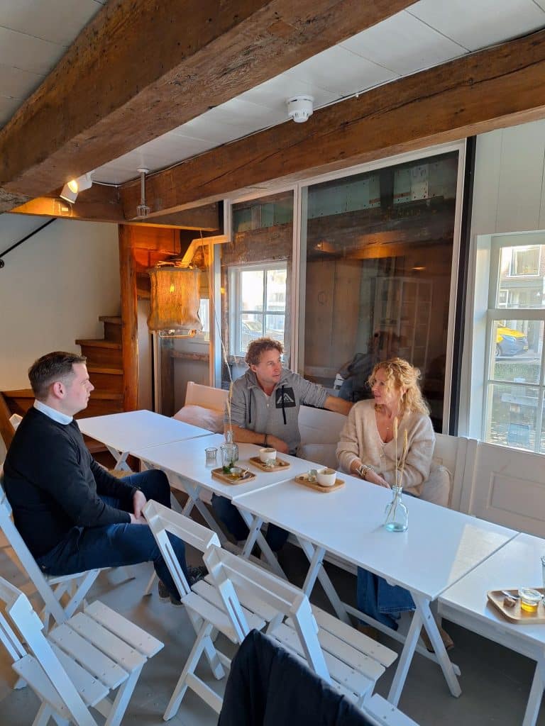Aan een lange houten tafel zitten van links naar rechts op de foto wethouder Christiaan Peetoom, Geert Net en Hester van Velzen te praten met een kop koffie