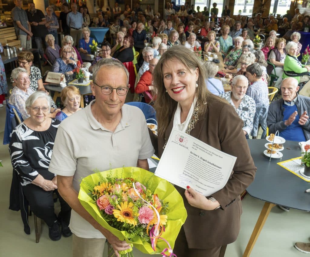 Engel Schoffelmeer met burgemeester Anja Schouten na uitreiking van het gouden Ere-insigne. Vrijwilligers van WonenPlus kijken toe