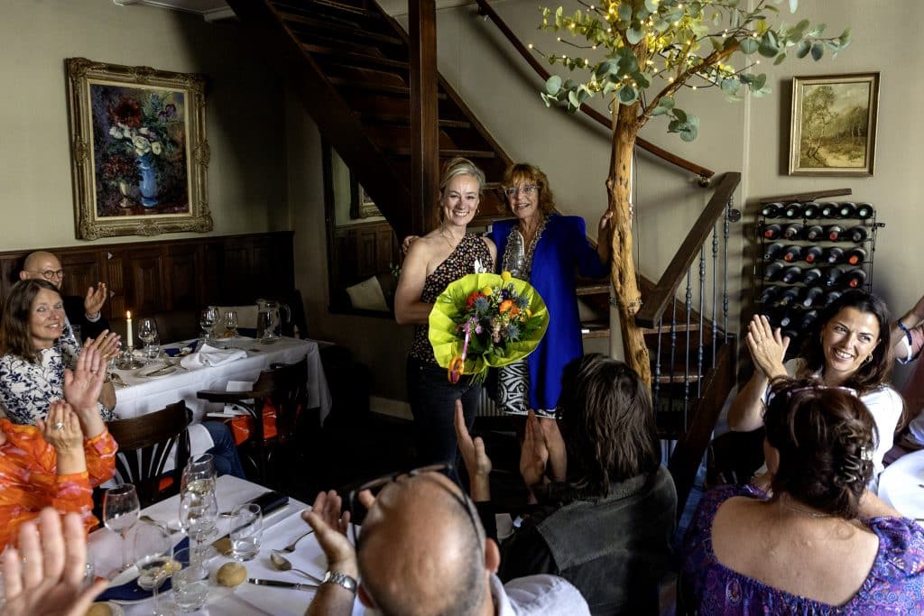 Judith Warries met bloemen naast locoburgemeester Van de Ven, omringd door genodigden in restaurant