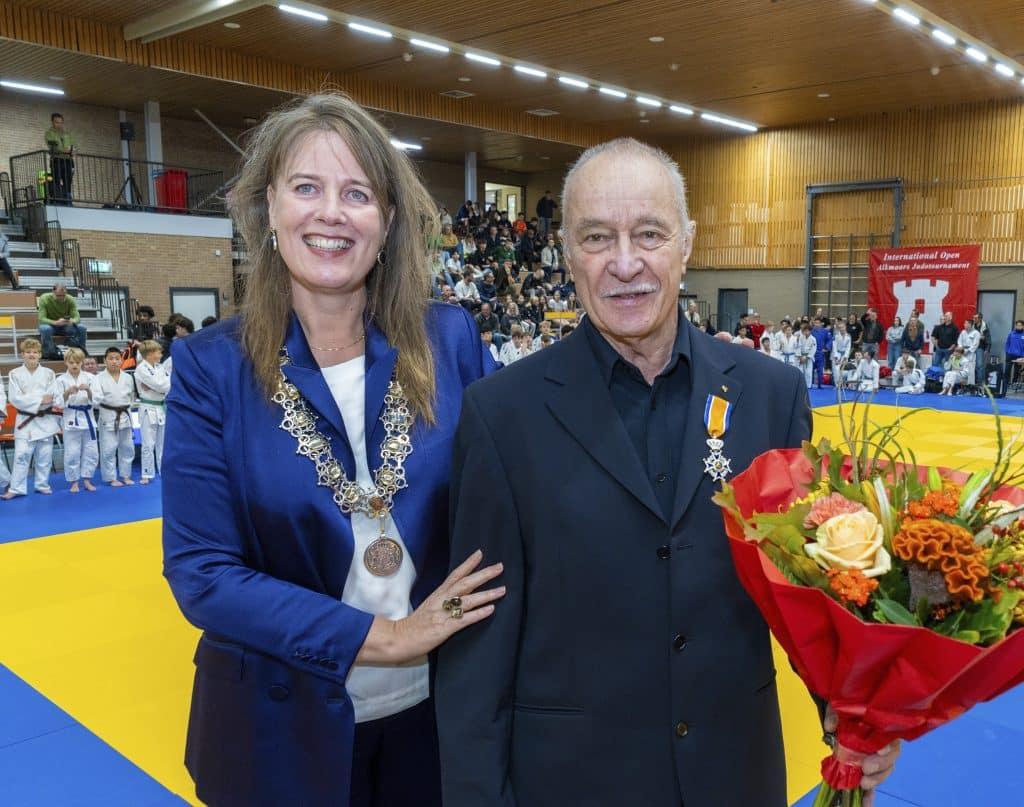 Burgemeester Anja Schouten met ambtsketen met rechts van haar judoka Ton Huijberts met bloemen en het zojuist opgespelde lintje. Op de achtergrond rond de tatami de deelnemers aan het Open Alkmaars judotoernooi. Fotograaf is Jan Jong