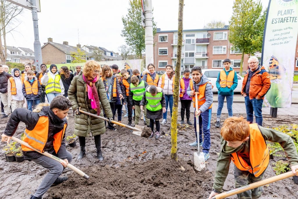 Foto van groep schoolkinderen en een volwassen vrouw met een schep. Samen planten ze een boom.