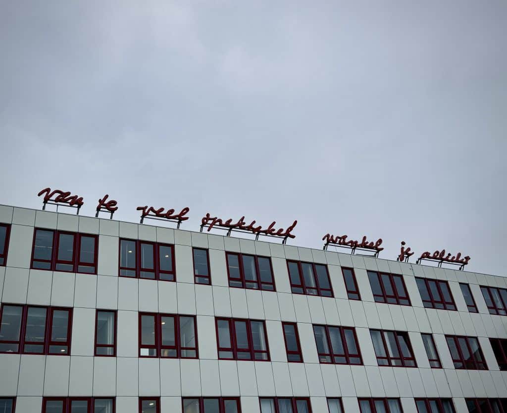 Foto van een wit gebouw met ramen met rode kozijnen. Het is het pand van het UWV in Alkmaar met daar bovenop het rode kunstwerk van Lucebert. De dichtregel "Van te veel spektakel wankel je allicht"