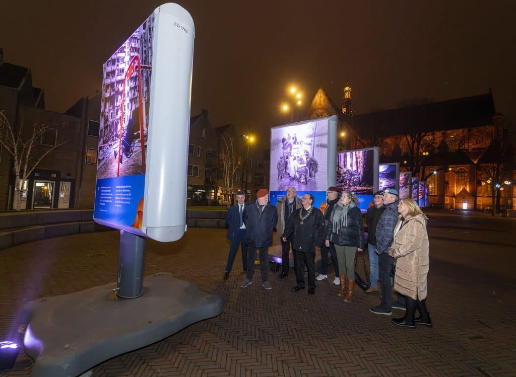 9 mensen staan naast elkaar op een plein en kijken naar een grote fototentoonstelling