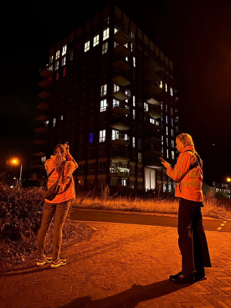 Twee vrouwen staan in het donker bij een flatgebouw met oranje hesjes. 