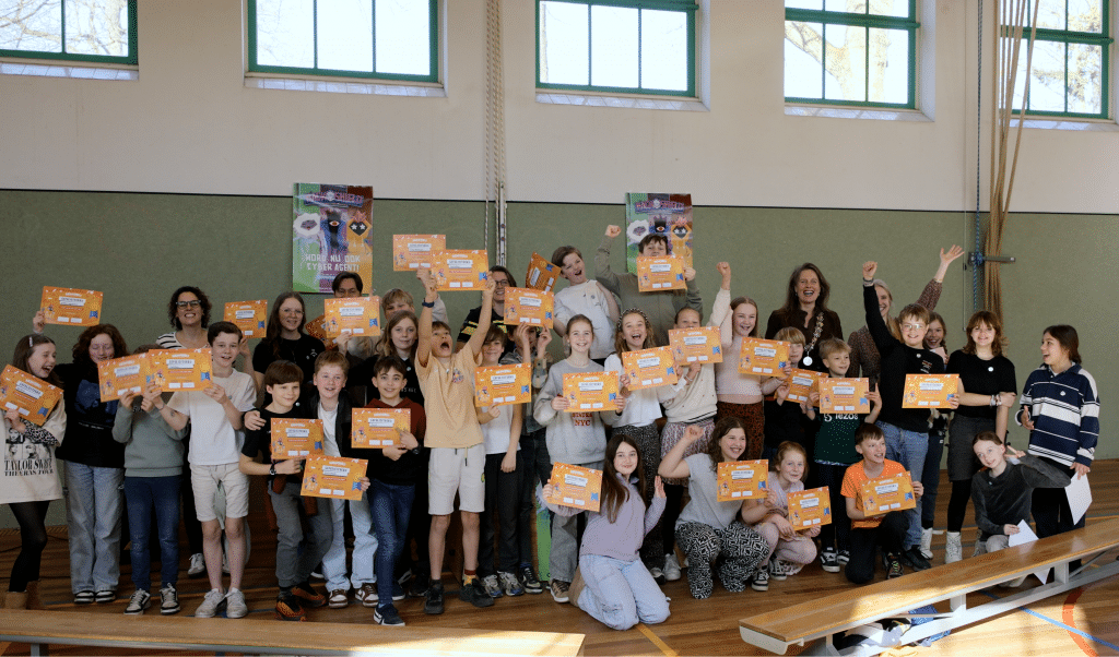 Een groep kinderen in een gymzaal. De kinderen juichen en houden een certificaat omhoog.