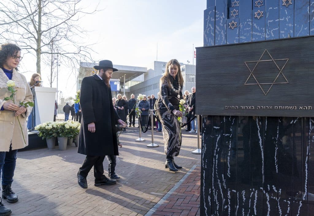 Drie mensen lopen richting een monument en leggen daar een witte roos neer. Op de achtergrond staat een groep mensen.