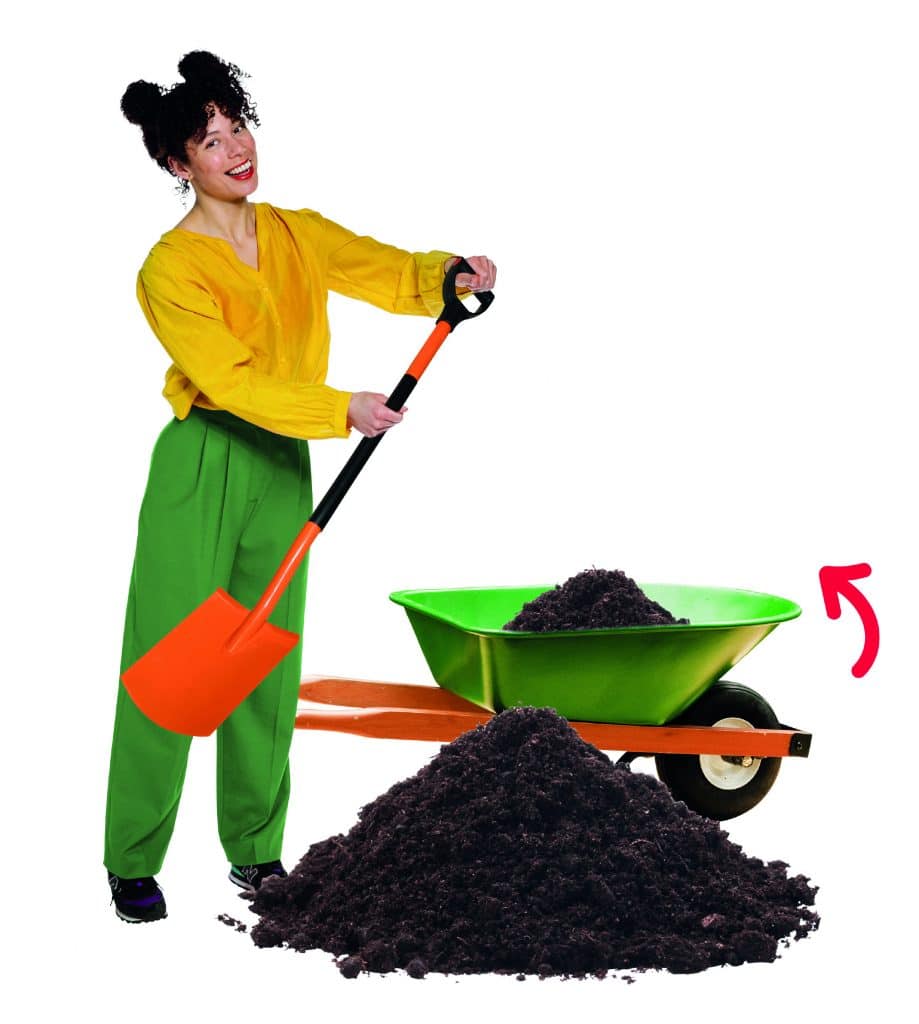 Vrouw met een schep in haar hand naast een hoop compost en een kruiwagen. 