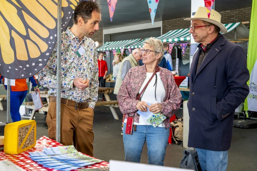 Drie mensen staan bij een marktkraam met een insectenhotel en zakjes met bloemzaad, een medewerker van de gemeente geeft uitleg. 