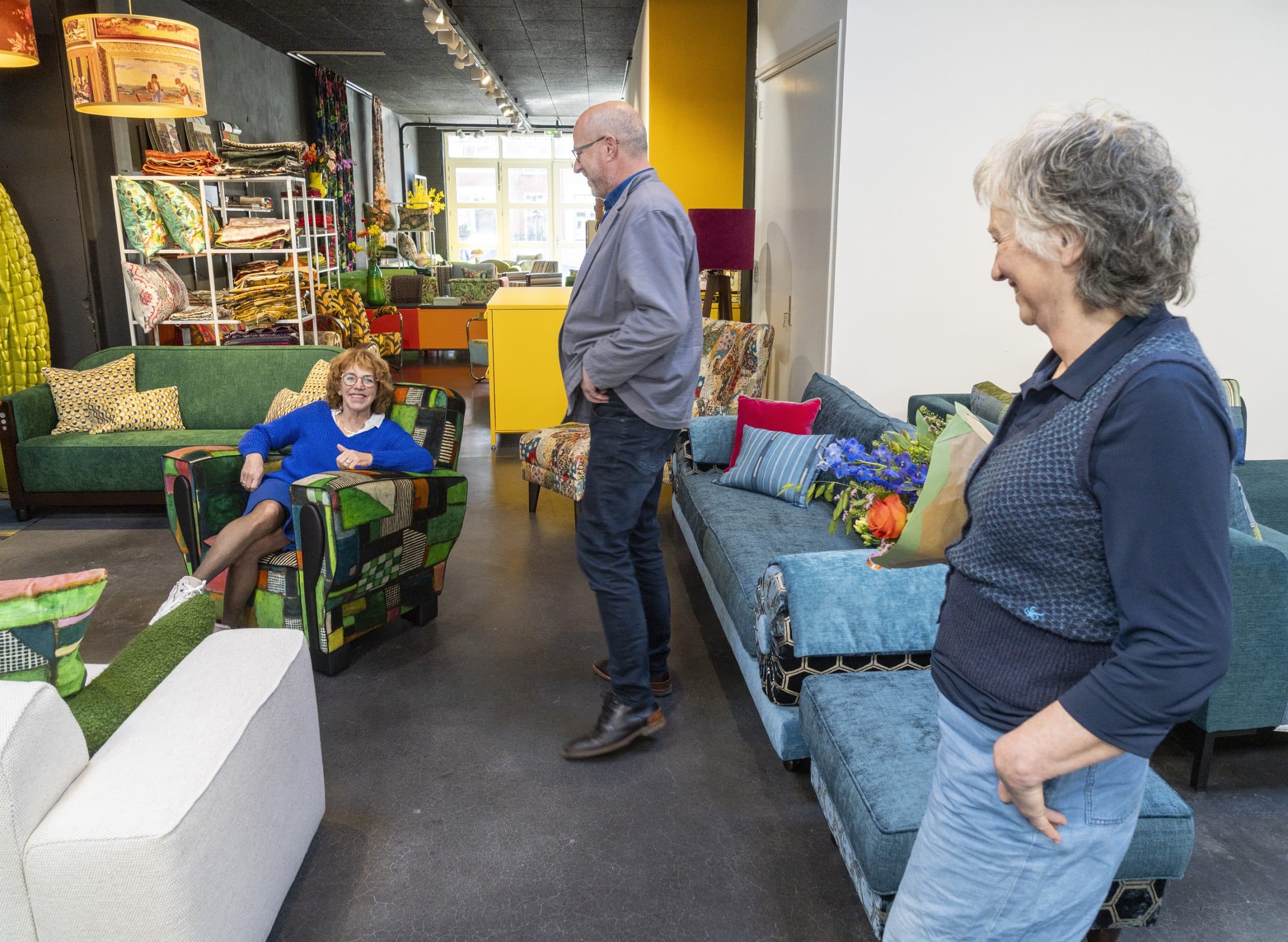 Wethouder Van de Ven in één van de fauteuils van Jantine Ranzijn. Jantine en haar partner Walter kijken haar lachend aan.