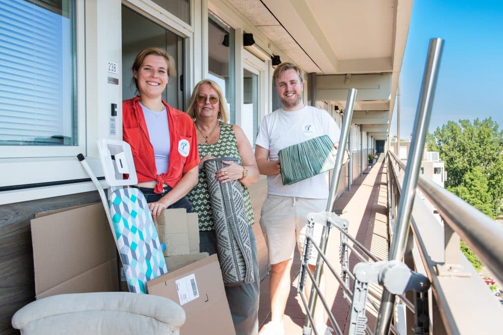 3 mensen op een galerij voor een appartement met spullen, zoals een strijkplank.