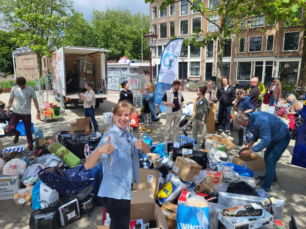 Vrouw steekt 2 duimen omhoog. Ze staat voor een plein dat vol ligt met spullen. Op de achtergrond een bestelbus. Er lopen en staan mensen die spullen inleveren of staan te praten.