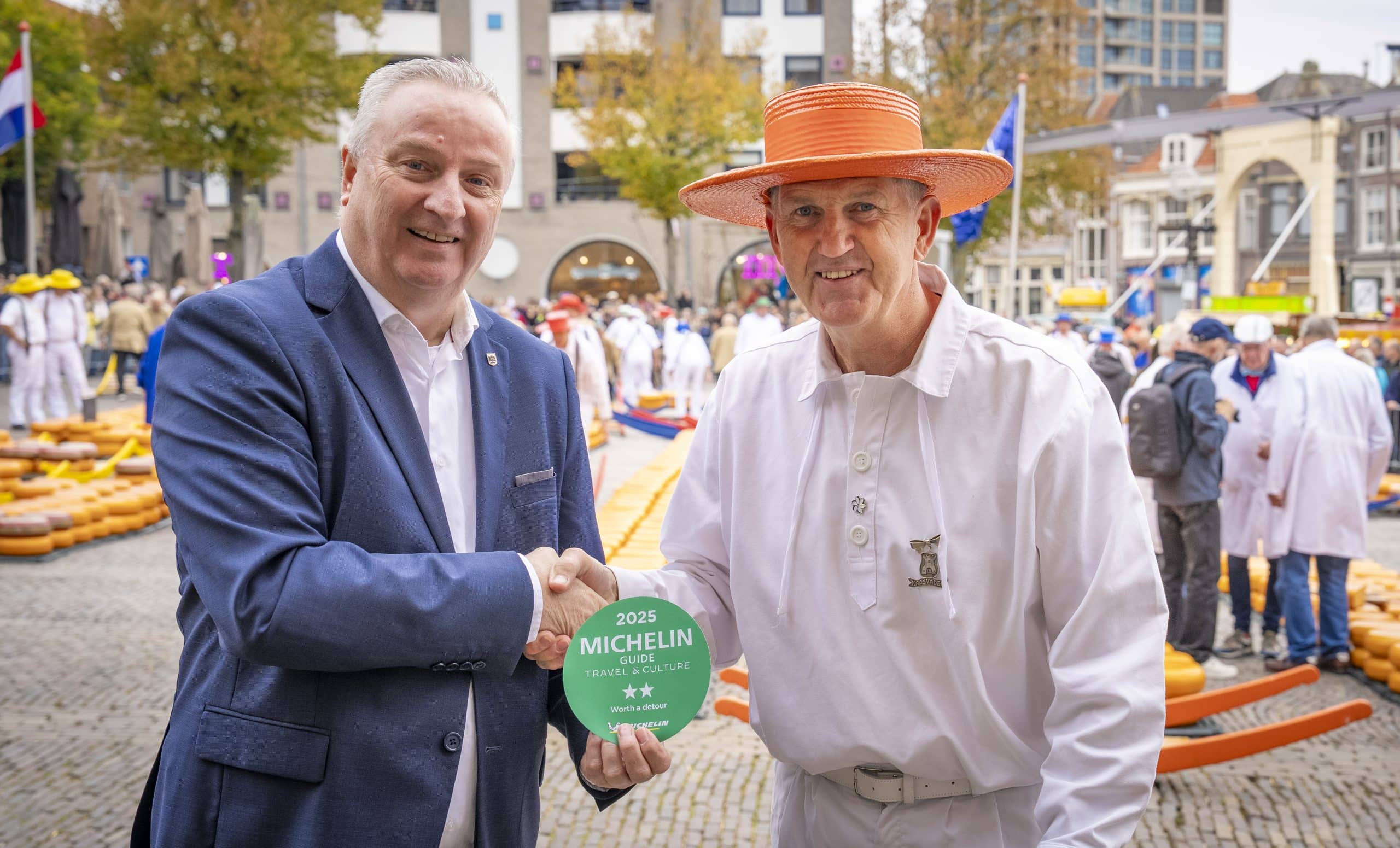 Wethouder Hoekzema geeft plaatsvervangend Kaasvader Harrie Konijn een hand en houdt de twee Michelinsterren voor de kaasmarkt voor in de vorm van een sticker. Op de achtergrond is de Alkmaarse kaasmarkt te zien