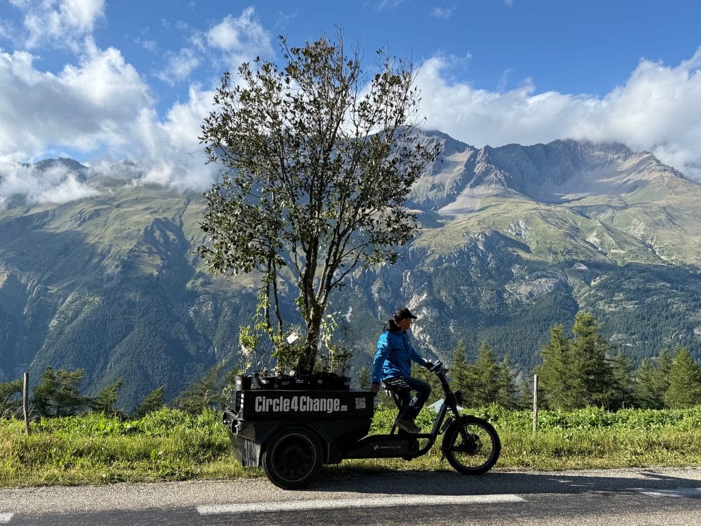 Bruno Doedens fietst op een fiets met een grote aanhanger met daarin een grote boom. Op de achtergrond bergen.