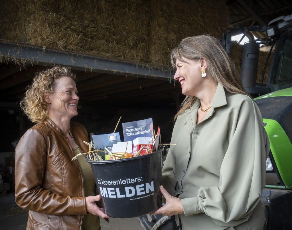 Burgemeester Schouten geeft een emmer met daarin producten, waaronder koffie, en een flyer aan een vrouw. Op de achtergrond staan hooibalen en een tractor.