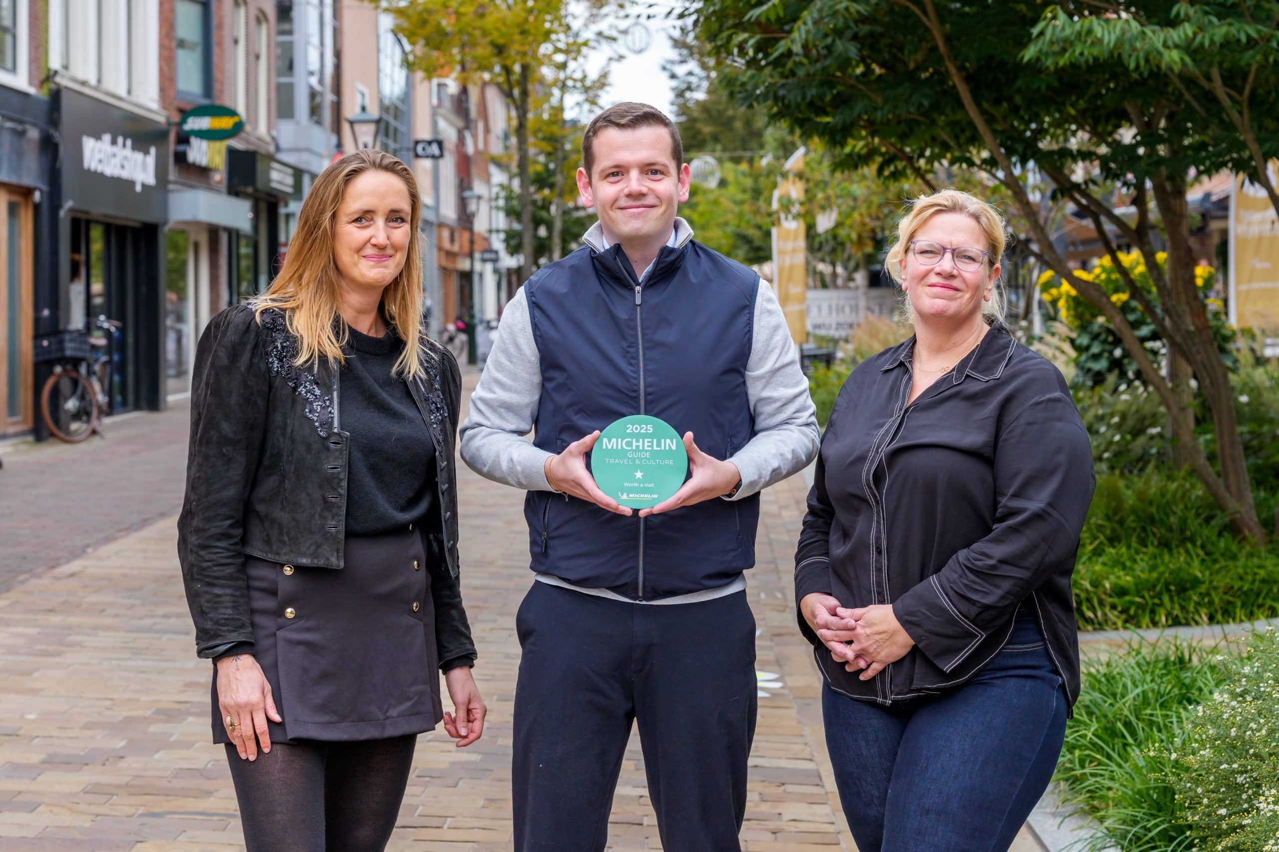 Wethouder Peetoom staat op de Laat in Alkmaar met twee Alkmaarse binnenstadondernemers naast hem aan beide kanten. Hij houdt de Michelinster voor de binnenstad vast.