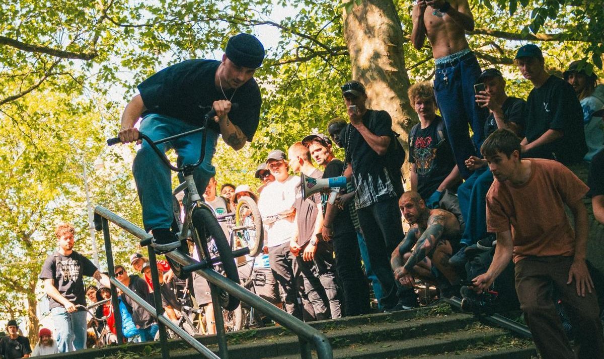 BMX'er gaat met fiets van een trapleuning af. Er staat een groep toeschouwers naar te kijken vanaf de zijlijn.