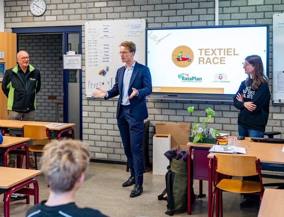 Wethouder Lars Ruiter in een klaslokaal met een docent en medewerker van RataPlan. Op de achtergrond een scherm met daarop een presentatie van de Textiel Race.