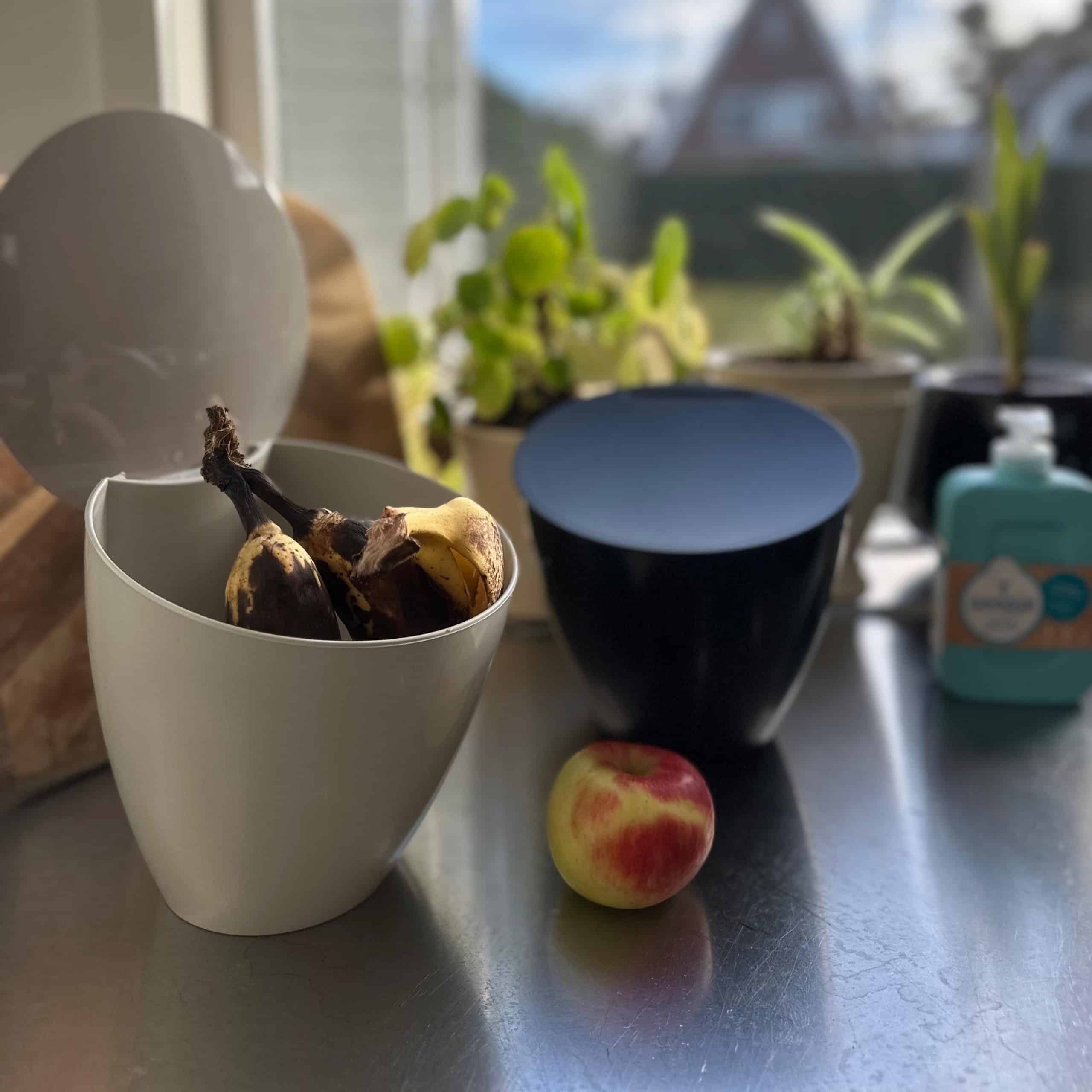Een zwart en wit gft-keukenbakje op het aanrecht. In het witte bakje zitten bananenschillen. Op het aanrecht ligt een appel.