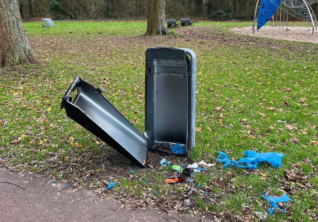 Een vuilnisbak in een groene omgeving. De voorkant van de vuilnisbak is beschadigd en hangt los. Vuilnis ligt op de grond bij de vuilnisbak