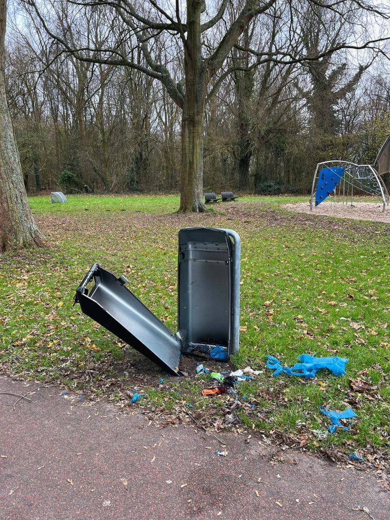 Een afvalbak in een groene omgeving. De voorkant van de afvalbak is beschadigd en hangt los. Vuilnis ligt op de grond bij de afvalbak 