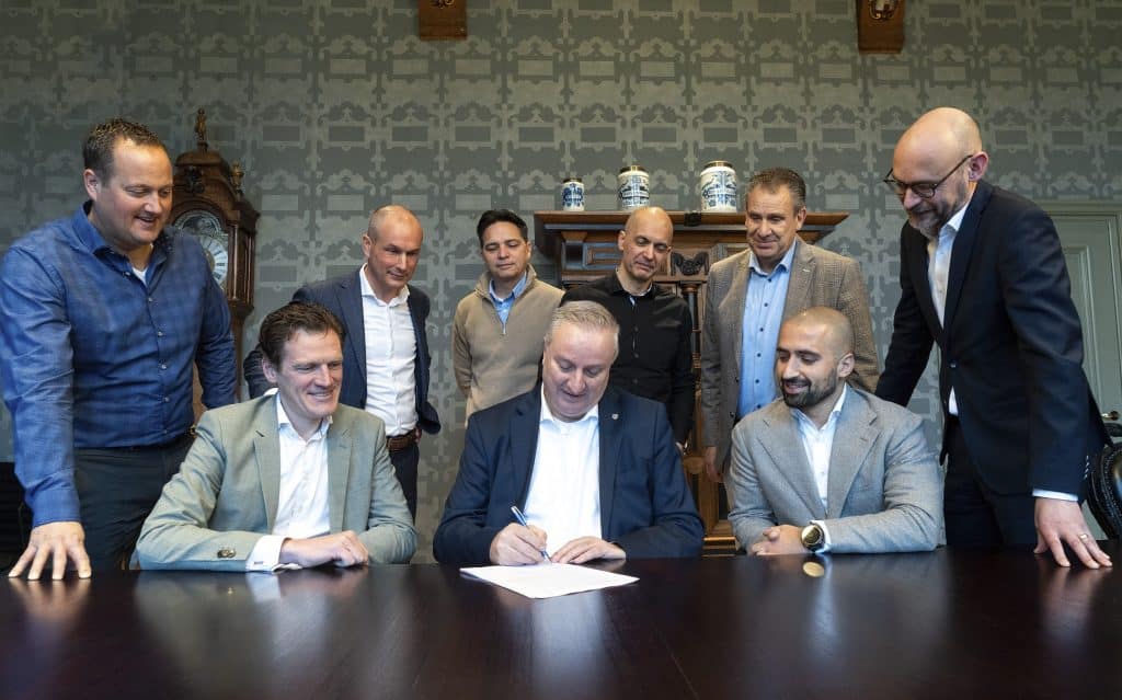 Groep mensen die achter een tafel zitten of staan en gezamenlijk naar een contract op tafel kijken. De man in het midden, de wethouder, ondertekent het contract.