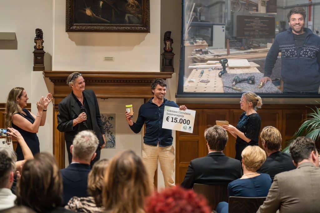 Man staat met voucher met daarop € 15.000 euro in zijn hand, naast hem klappende mensen. Op de achtergrond zijn foto op het scherm.