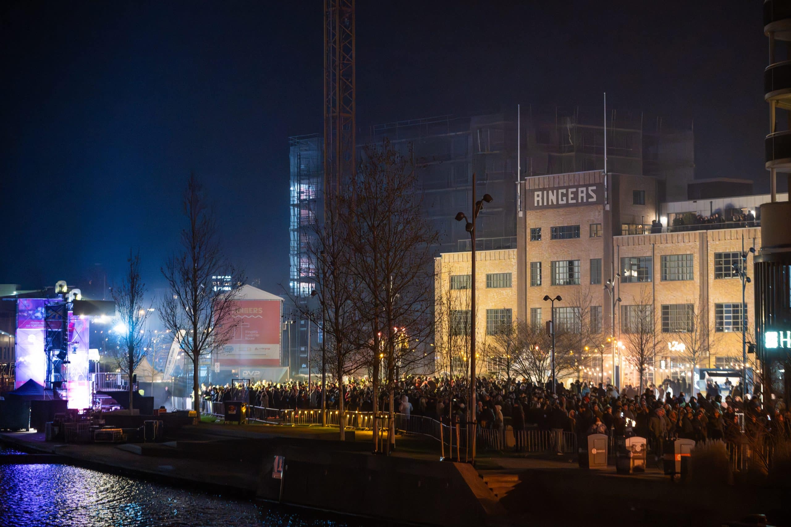Een grote groep mensen staat op een plein en kijkt naar een podium met licht en lasers.