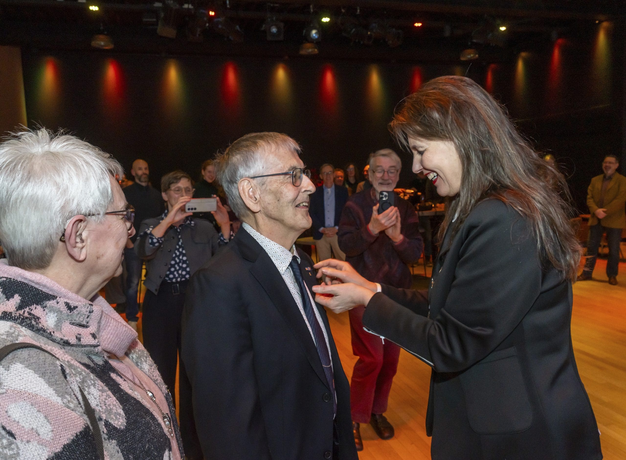 In een theaterzaal staat op de voorgrond een ouder echtpaar samen met burgemeester Anja Schouten. Bij de man wordt her ere-insigne van de gemeente Alkmaar opgespeld door de burgemeester. Op de achtergrond kijken mensen toe en ze maken foto's.