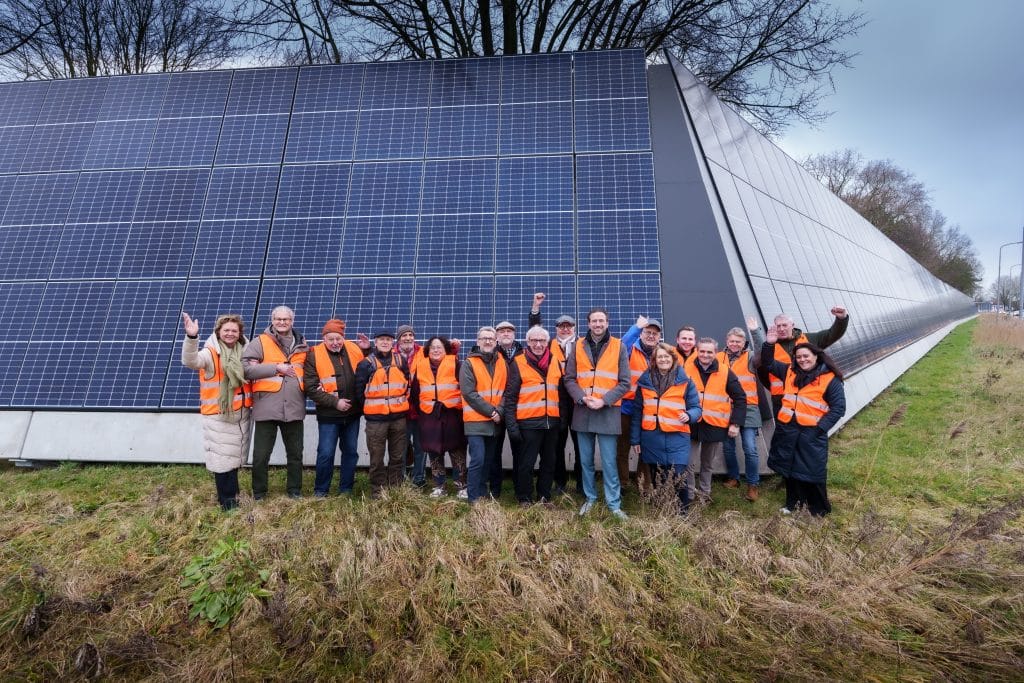 Groep bewoners en betrokkenen in oranje veiligheidshesjes voor een groot geluidsscherm met zonnepanelen langs een weg in Alkmaar.
