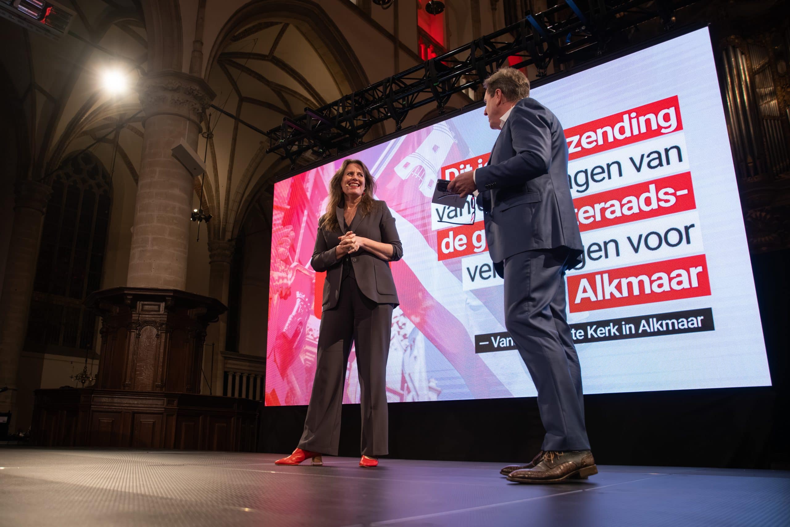 Een man en een vrouw staan op een podium voor een scherm en lachen naar elkaar.