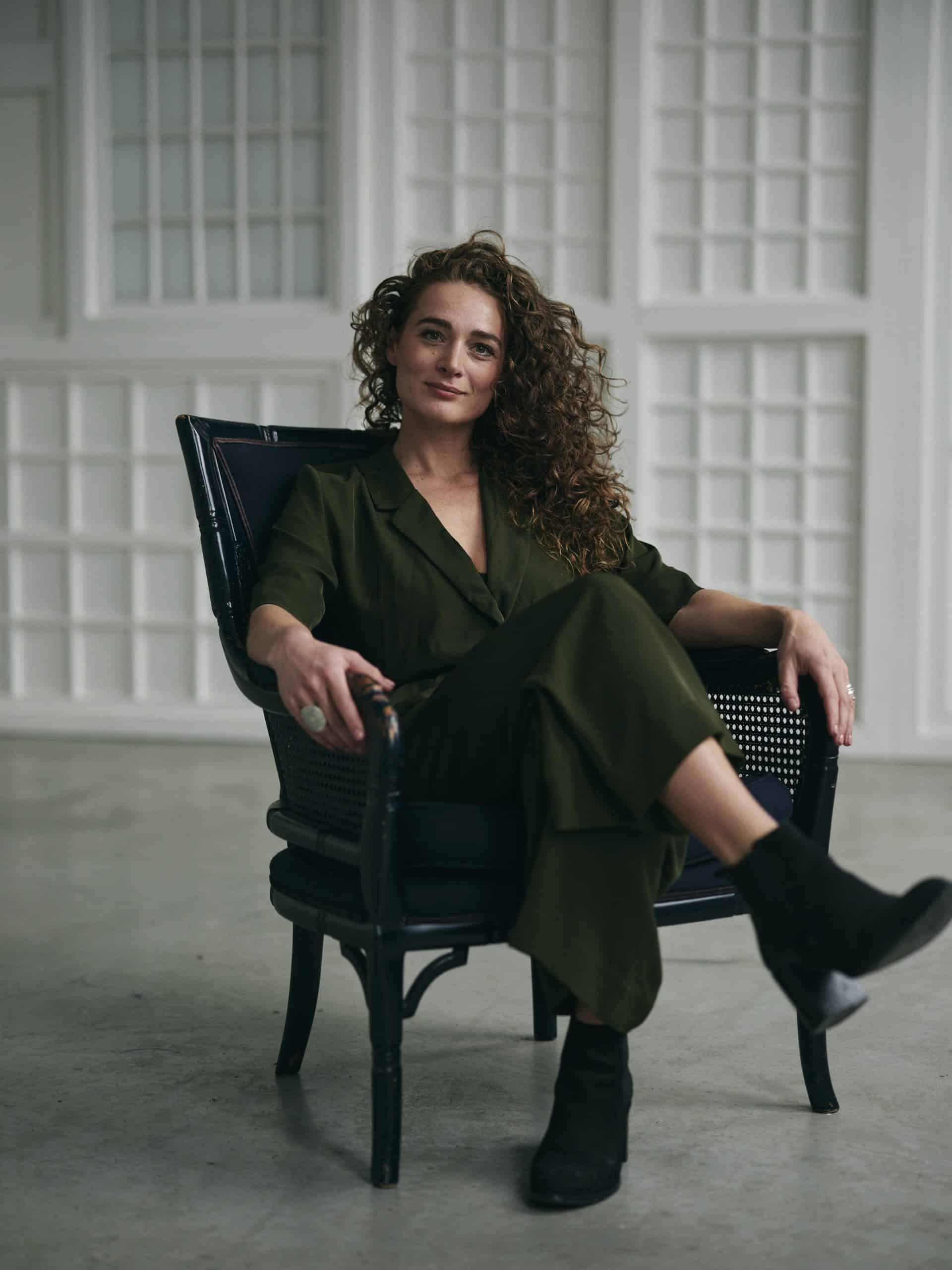 Een vrouw met rode krullen en een groen pak zit in een stoel