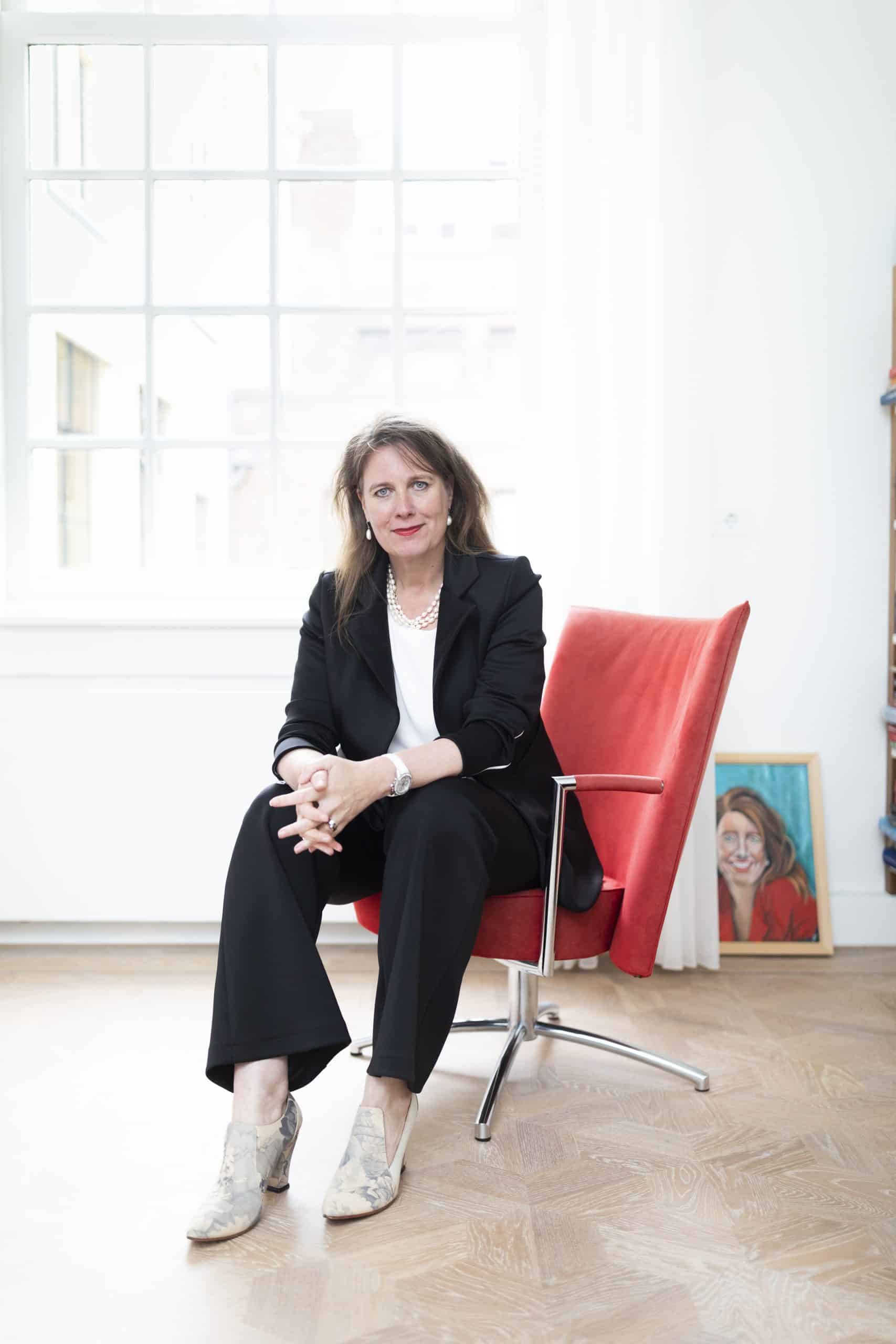 Een vrouw in zwart pak zit op een rode stoel en kijkt vriendelijk