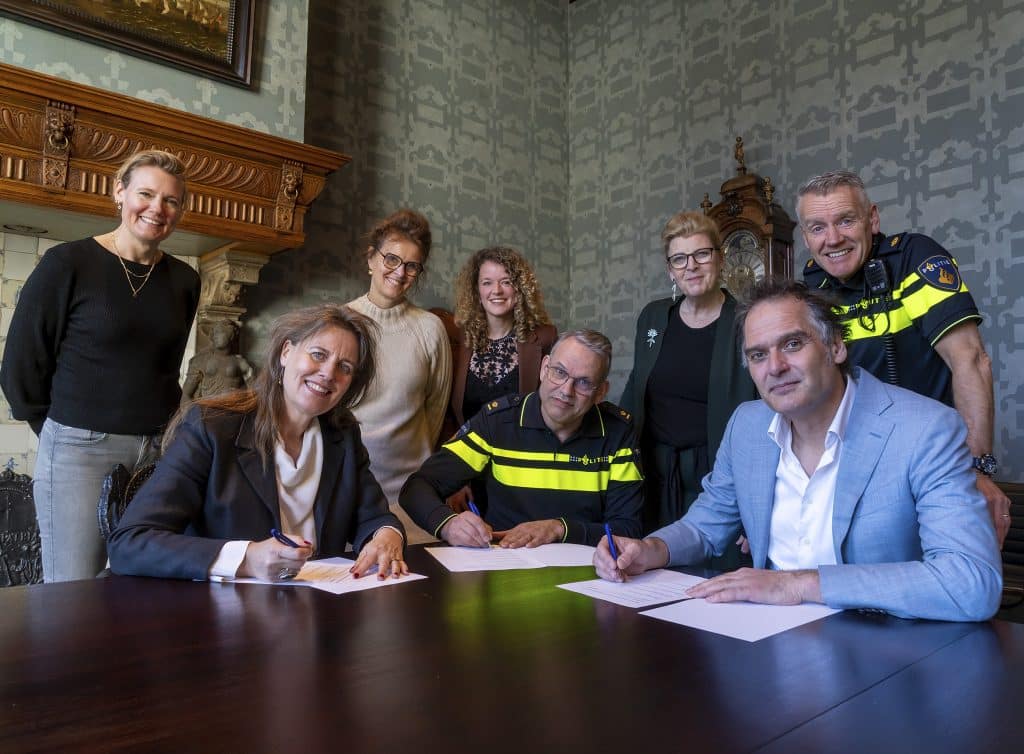 Drie mensen zitten aan een bureau en ondertekenen een document. Daarachter staan 5 mensen.