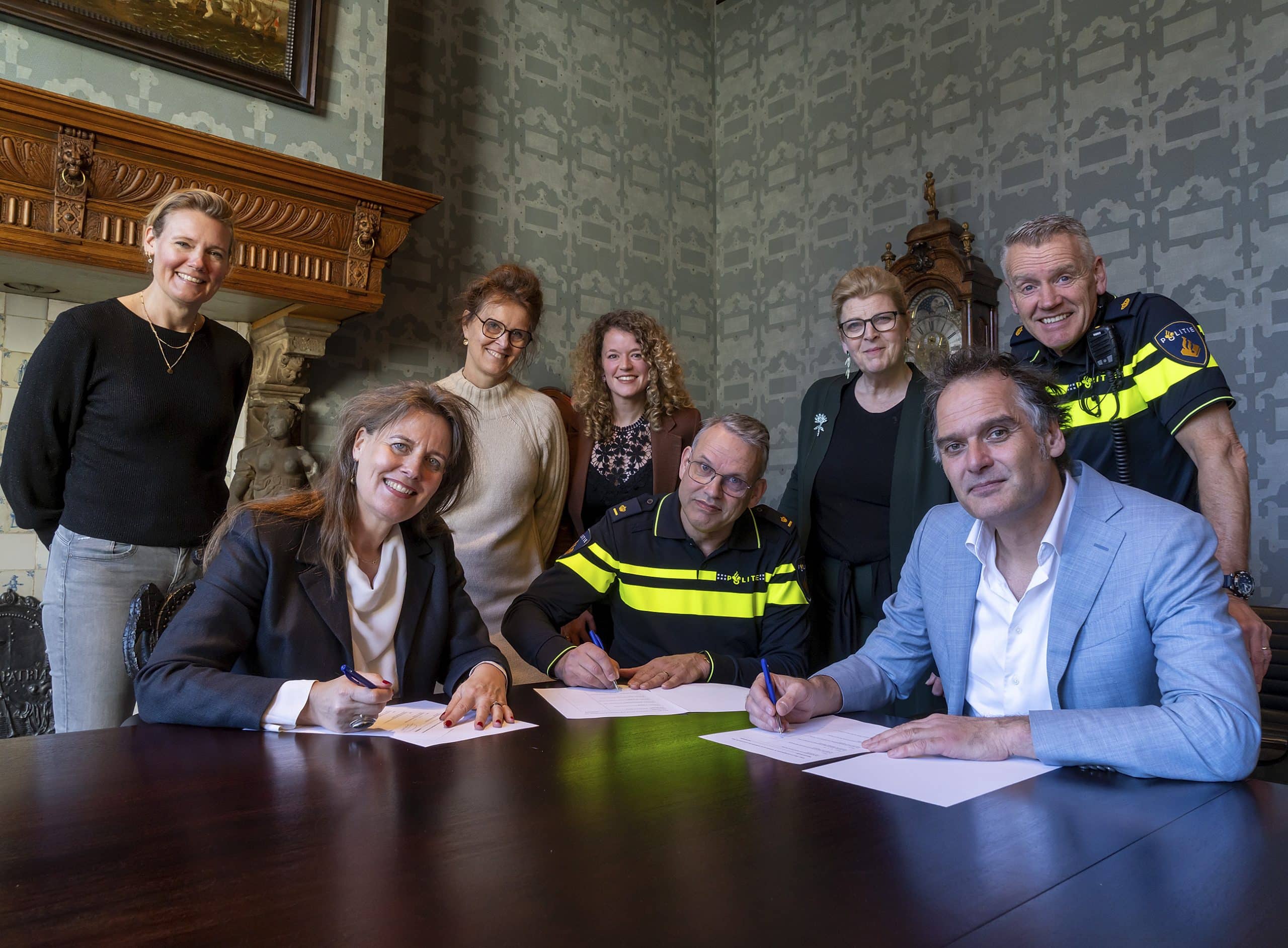 Drie mensen zitten aan een bureau en ondertekenen een document. Daarachter staan 5 mensen.