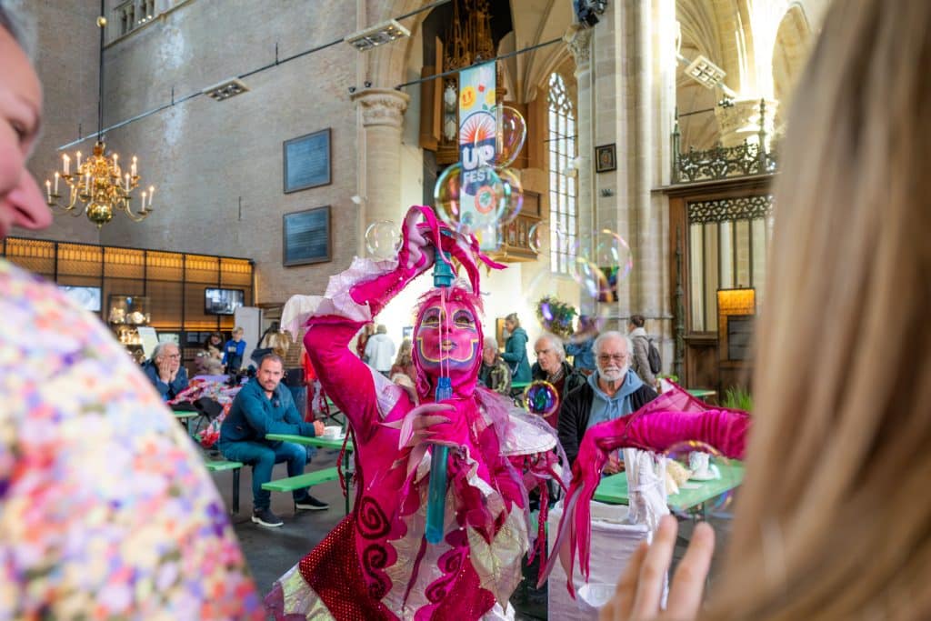 Artiest in een roze fluwelen pak met roze make up. Houdt een grote bellenblaas vast en blaas grote bellen. Er staan mensen omheen. Het is binnen in de Grote Kerk Alkmaar.