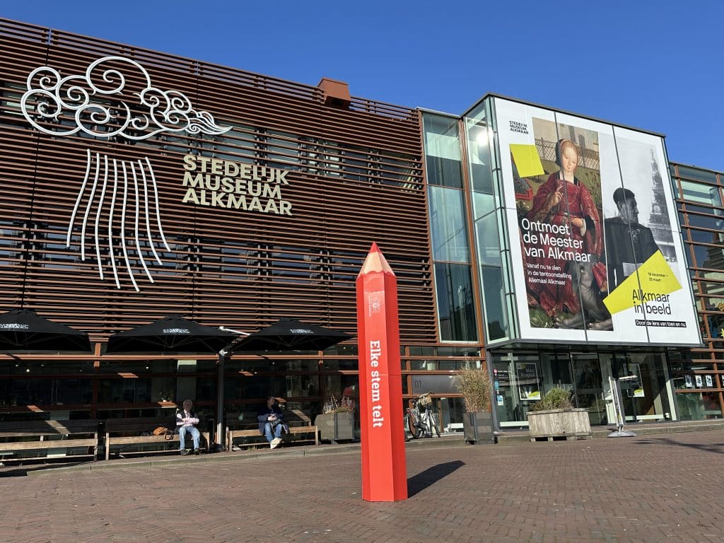 Op een plein staat een groot rood potlood met de tekst "Elke stem telt". Op de achtergrond het Stedelijk Museum Alkmaar