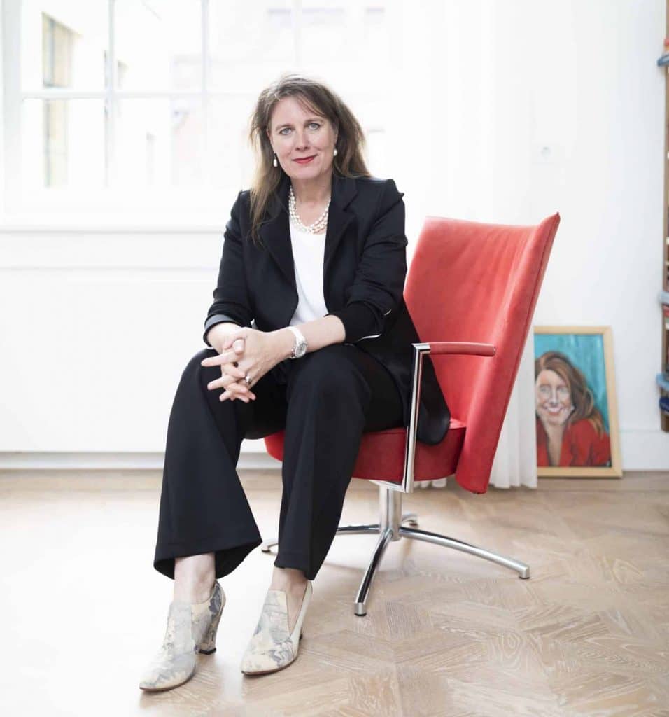 Burgemeester Anja Schouten zit op een rode stoel en kijkt in de camera.